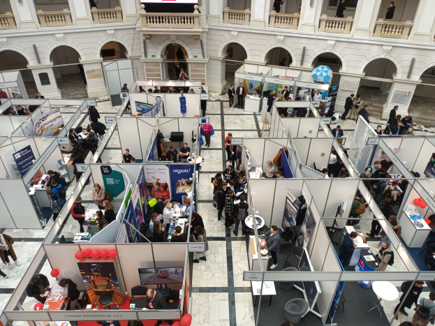 Eine große Gruppe von Menschen versammelte sich in einem Raum voller Stände, Banner, Tische, Stühle und Ballons, mit einem Gebäude im Hintergrund, das Bögen, Säulen, Geländer und einen Bildschirm aufweist.