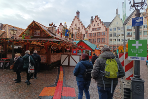 Eine Gruppe von Menschen geht eine Kopfsteinpflasterstraße neben einem Weihnachtsmarkt in Nürnberg, Deutschland, entlang, wobei einige Taschen, Laternenmasten und Texttafeln zu sehen sind und Gebäude mit Fenstern und ein bewölkter Himmel im Hintergrund.