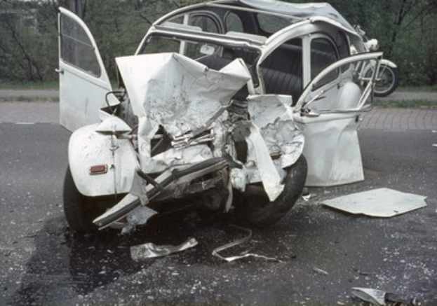Ein schwer beschädigtes Auto mit Frontalaufprall am Straßenrand, umgeben von Bäumen und einem Gebäude, mit einem weiteren Fahrzeug im Hintergrund.