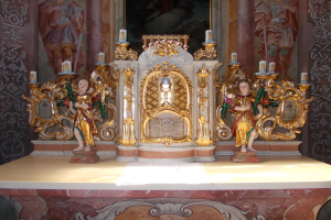 Ein Altar in einer Kirche mit Skulpturen von Menschen und Kerzen auf Ständern, vor einer Wand mit einem Gemälde.