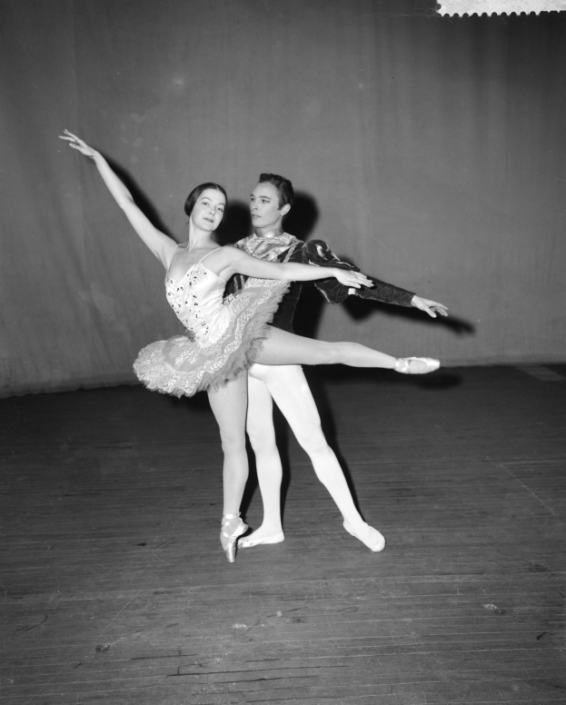 Ein Schwarz-Weiß-Foto eines Mannes in weißem Hemd und schwarzen Hosen und einer Frau in unbekannter Kleidung, die perfekt synchron auf einer Bühne tanzen, mit einem Vorhang im Hintergrund.