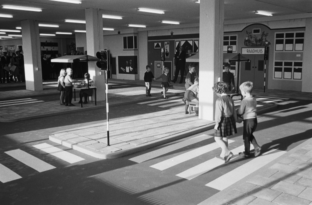 Schwarzes und weißes Foto von Kindern, die eine Straße an einer Ampel überqueren, mit Menschen auf Bänken, einem Verkehrszeichen und Hintergrundsäulen mit Deckenleuchten.