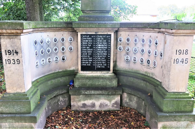 Ein Holocaust-Gedenkmonument in einem jüdischen Friedhof in Berlin, mit einer Tafel mit Text und Zahlen, umgeben von Bäumen, einem Zaun und verstreuten trockenen Blättern.
