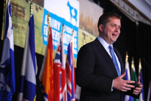 Ein Mann im Anzug und Krawatte steht an einem Rednerpult mit Mikrofon, umgeben von Flaggen und einem Hintergrundbild.