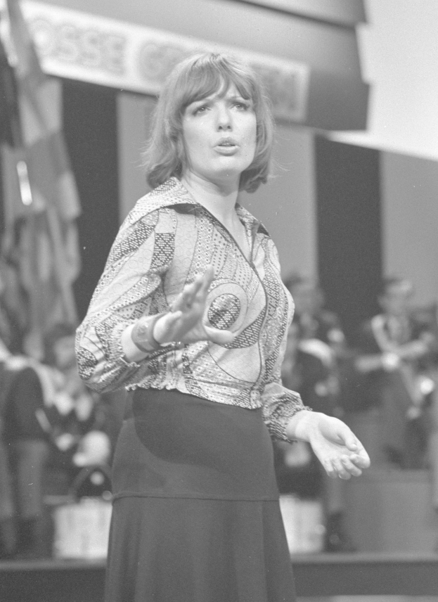 Eine Frau in einem Schwarz-Weiß-Foto steht auf einer Bühne, trägt ein weißes Hemd und einen schwarzen Rock, hat ihre Haare zu einem Knoten gebunden und einen ernsten Gesichtsausdruck, umgeben von einer Gruppe von Menschen und einer Wand im Hintergrund.