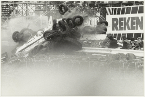 Ein Schwarz-Weiß-Foto eines zertrümmerten Rennwagens, umgeben von Reifen, mit einer Gruppe von Menschen auf der rechten Seite und einer Tafel mit Text im Hintergrund.