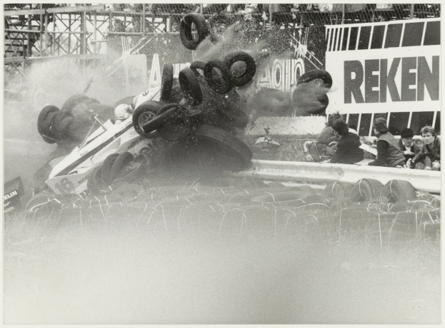 Ein Schwarz-Weiß-Foto eines zertrümmerten Rennwagens, umgeben von Reifen, mit einer Gruppe von Menschen auf der rechten Seite und einer Tafel mit Text im Hintergrund.