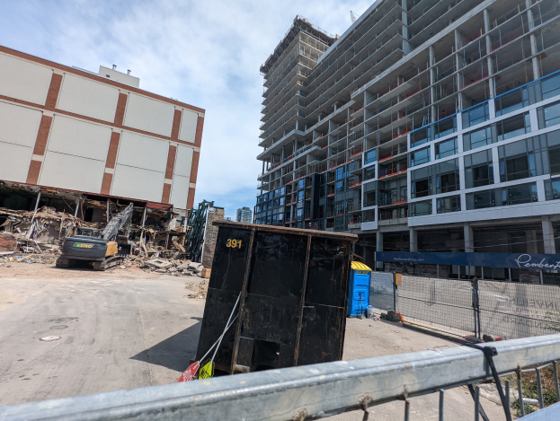 Großes Gebäude mit viel Schutt davor, umgeben von einem Metallzaun, einem Container, Pflanzen, Bäumen, Pfählen, einem Schild, Fahrzeugen, Steinen und einem bewölkten Himmel, was auf eine Baustelle für einen neuen Wohnkomplex hinweist.