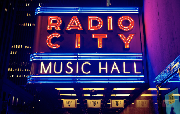 Außensicht des Radio City Music Hall in New York City bei Nacht, hell erleuchtet mit hohen Gebäuden im Hintergrund, mit einer 'Radio City'-Leuchtschrift und mehreren kleineren Leuchtschriften auf der rechten Seite.