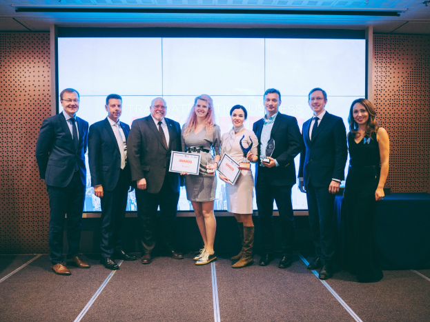 Gruppe strahlender Menschen mit Urkunden, die vor einem Bildschirm mit einem Tisch und einem Wandobjekt im Hintergrund stehen, Gewinner der International Business Awards 2018.