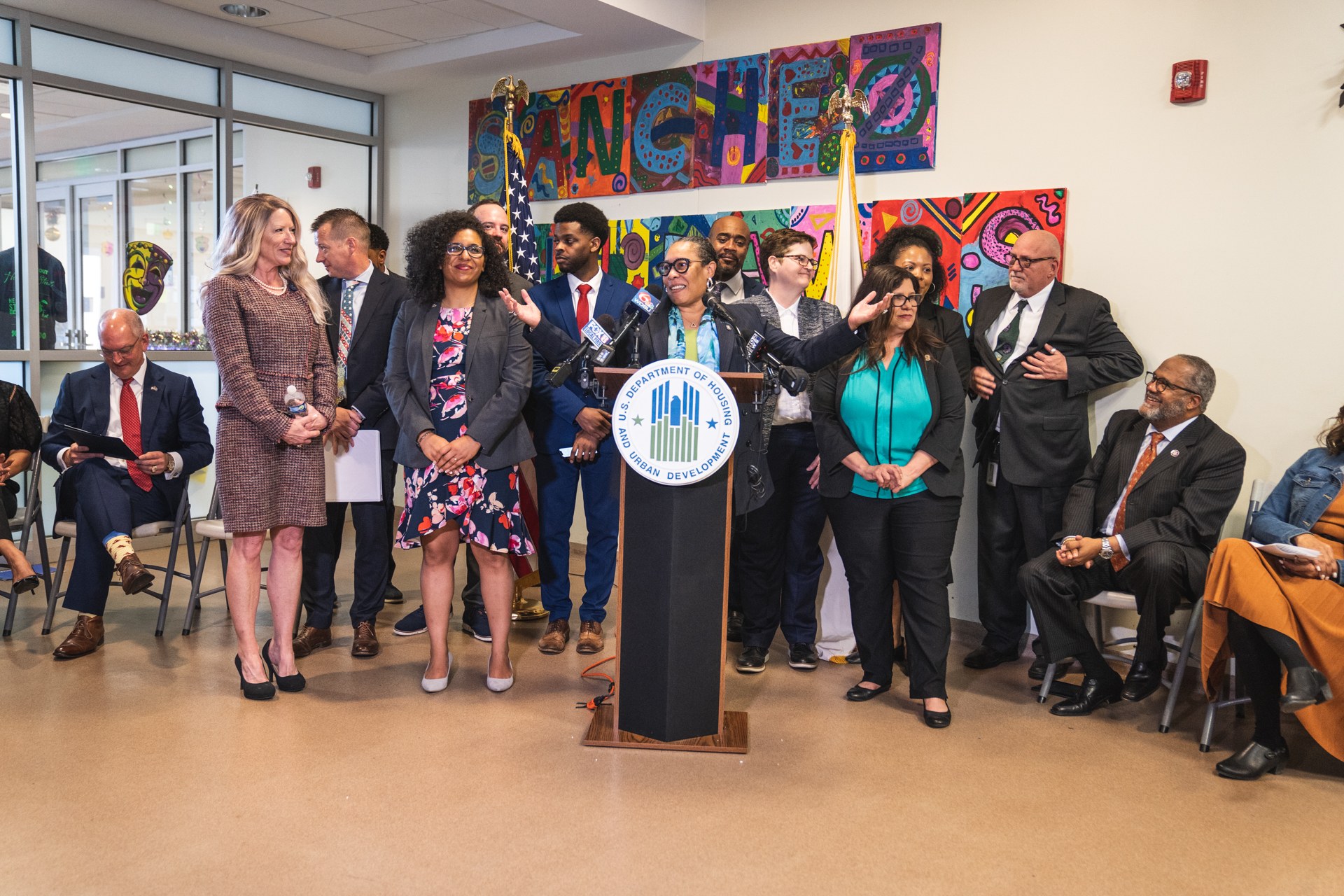 Gruppe von Menschen vor einem Rednerpult mit Mikrofon und einer Tafel mit der Aufschrift "Sancho-Stadtrat tritt dem Stadtrat bei", mit sitzenden und stehenden Zuschauern, Flaggen, einem Feuerlöscher und Deckenleuchten im Hintergrund.