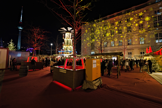Ein lebendiger Weihnachtsmarkt in Berlin, Deutschland, mit Menschen um beleuchtete Stände, Tannen, Gebäude, Laternenmäste und einen Turm unter einem dunklen Himmel.