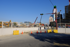 Eine Baustelle mit zahlreichen Verkehrskegeln, eine Person auf der Straße umgeben von Absperrungen, ein Zaun, ein Schild, ein Straßenschild, eine Ampel, zusätzliche Pfosten, Gebäude mit Fenstern, Kräne, Bäume und ein bewölkter Himmel.