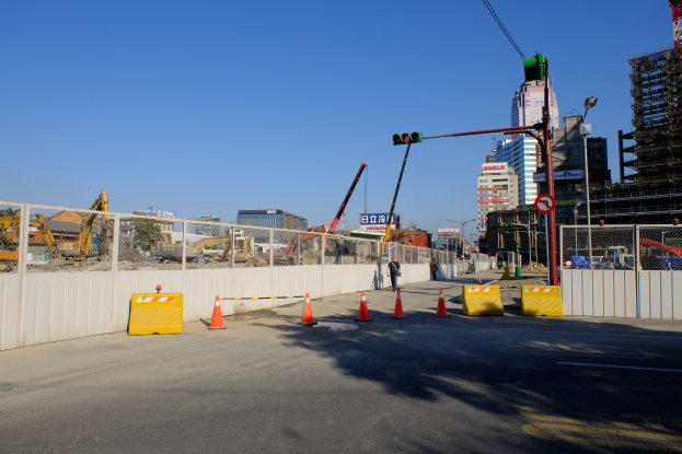 Eine Baustelle mit zahlreichen Verkehrskegeln, eine Person auf der Straße umgeben von Absperrungen, ein Zaun, ein Schild, ein Straßenschild, eine Ampel, zusätzliche Pfosten, Gebäude mit Fenstern, Kräne, Bäume und ein bewölkter Himmel.