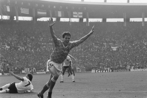 Ein Schwarz-Weiß-Foto eines Fussballspielers, der nach einem Tor feiert, umgeben von Menschen auf dem Boden, mit Schildern, Fahnen und Pfählen im Hintergrund unter einem klaren Himmel.