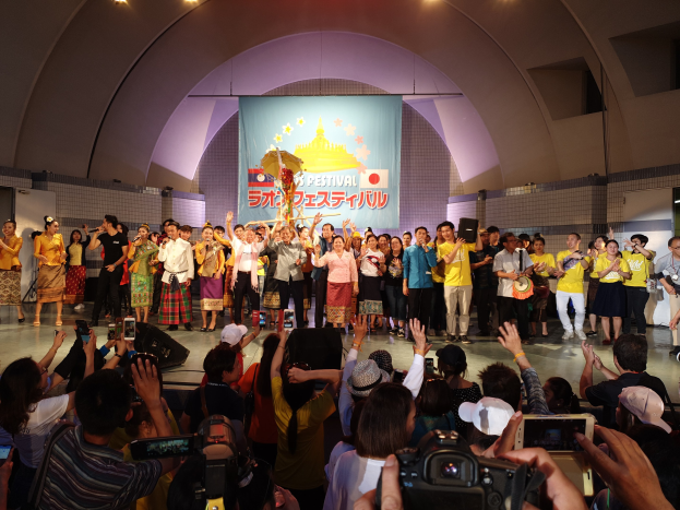 Eine Gruppe von Menschen auf einer Bühne mit Kameras, Handys und Musikinstrumenten während einer Festival-Eröffnung, mit einem Banner, Wänden und Deckenlampen im Hintergrund.