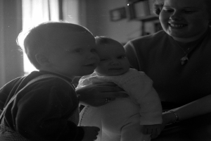 Eine Frau hält ein Baby mit einem warmen Lächeln im Arm, während zwei andere Kinder neben ihr stehen, in einem gemütlichen Raum mit Rahmen an der Wand und einem Fenster im Hintergrund.
