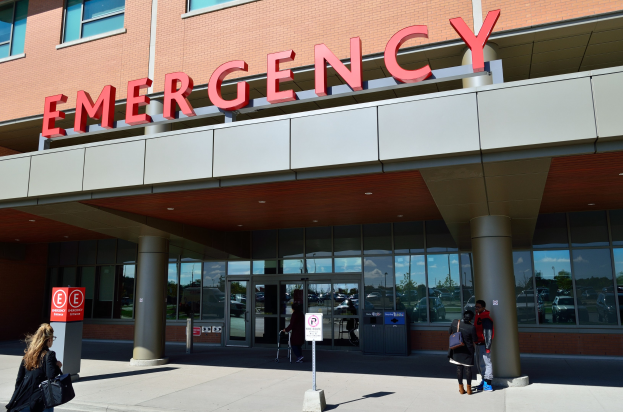 Eine Gruppe von Menschen geht vor einem Glaswand-Notfallgebäude mit Säulen, einem Namensschild, Hinweistafeln auf Pfählen und einem Abfalleimer vorbei.