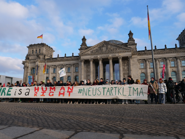 Eine Gruppe von Menschen mit einem Banner mit der Aufschrift "Wir sind ein Menschenrecht" vor dem Reichstaggebäude in Berlin, Deutschland, mit seinen architektonischen Details und umgeben von Fahnen unter einem bewölkten Himmel.