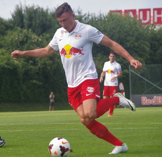 Ein professioneller Fußballspieler schießt einen Ball auf einem grünen Feld mit Bäumen und einem klaren blauen Himmel im Hintergrund, wobei ein Schild mit der Aufschrift "RB Leipzig" sichtbar ist.