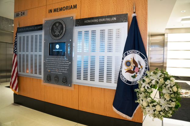 Gedenktafel in einer Lobby mit Flaggen, einem Kränzchen, einem Blumenstraß im Vordergrund, einer Wand mit Brettern und Text, Deckenleuchten und Aufzügen auf der linken Seite.