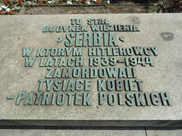 Gedenkstein mit Inschrifttafel vor einem Gebäude umgeben von blühenden Pflanzen.