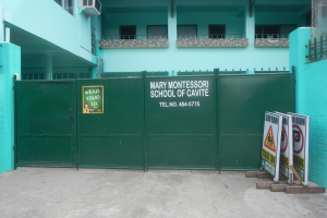 Mary Montessori School of Cavite, ein mehrstöckiges Gebäude mit Fenstern, Geländern, Säulen und Klimaanlagen, das ein grünes Tor mit einem Schild und Tafeln mit Text auf der rechten Seite zeigt.