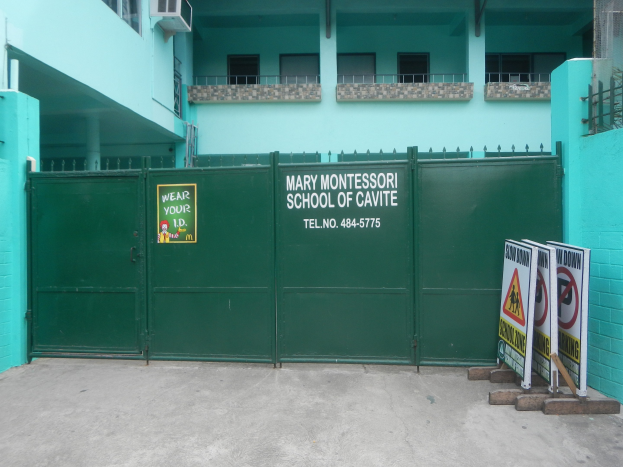 Mary Montessori School of Cavite, ein mehrstöckiges Gebäude mit Fenstern, Geländern, Säulen und Klimaanlagen, das ein grünes Tor mit einem Schild und Tafeln mit Text auf der rechten Seite zeigt.