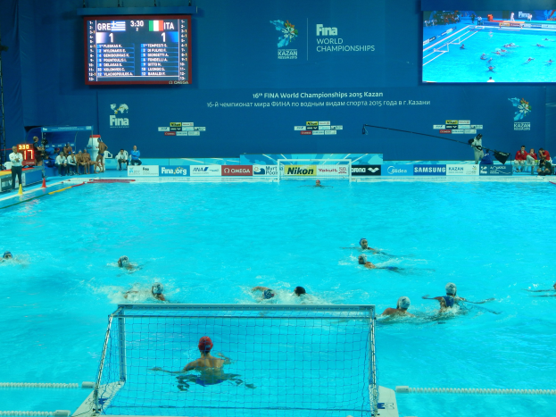 Eine Gruppe von Menschen spielt Wasserball in einem großen Pool mit einem Netz in der Mitte, Zuschauern auf der linken Seite und einer Wand im Hintergrund mit Text und Logos für die FINA-Weltmeisterschaften 2016 in Kazan.
