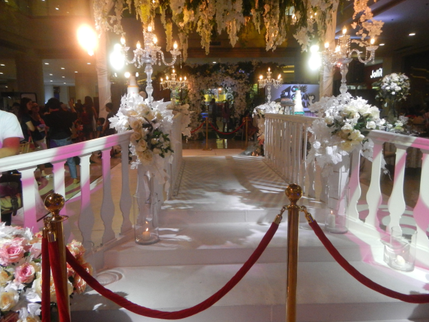 Eine Hochzeitszeremonie in einem Einkaufszentrum mit weißem Teppich, dekorativen Geländern, Blumenarrangements, Kronleuchtern und Deckenbeleuchtung, bei der eine Menschenmenge anwesend ist, darunter einige mit Taschen.