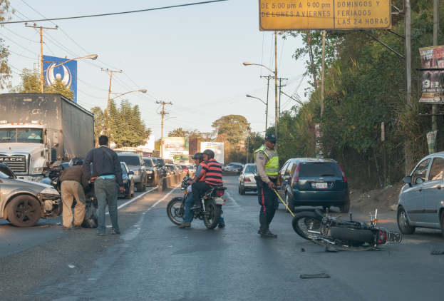 Eine Gruppe von Menschen steht um ein verunglücktes Motorrad auf der Seite einer Straße mit mehreren Fahrzeugen, darunter ein Lastwagen, und einem Hintergrund aus Bäumen, Pfählen, Lampen und Schildern.