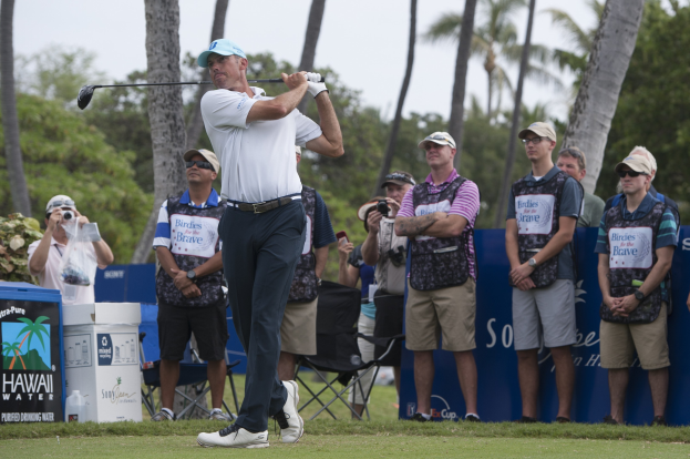 Ein Mann in einem weißen T-Shirt, schwarzen Hosen und einer blauen Mütze schwingt einen Golfschläger auf einem saftig grünen Feld, während eine Gruppe von Menschen mit Kameras zusieht, mit Bäumen und einem klaren blauen Himmel im Hintergrund.