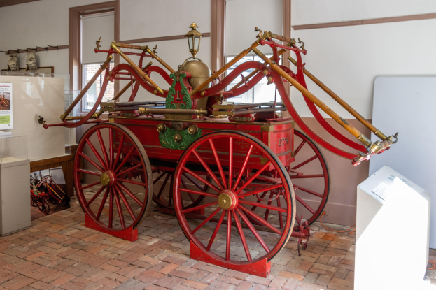 Altes Feuerwehrauto in einem Museum ausgestellt, umgeben von Barrieren und Ausstellungsstücken, mit Fenstern und einer Wand im Hintergrund.