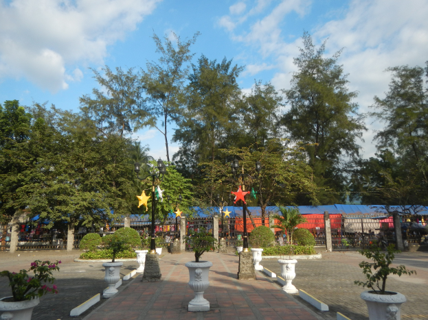 Ein Park mit Topfpflanzen und Bäumen, umgeben von Straßenlaternen, Schmuckelementen und Grills, mit einem bewölkten Himmel im Hintergrund.