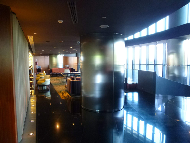 Eine geräumige Business-Lounge am Hong Kong International Airport mit großen Fenstern, hohen Decken mit elektrischem Licht, Sitzgruppen einschließlich Stühle und Sofas, Tischen, einem Teppichboden und einer zentralen Säule.