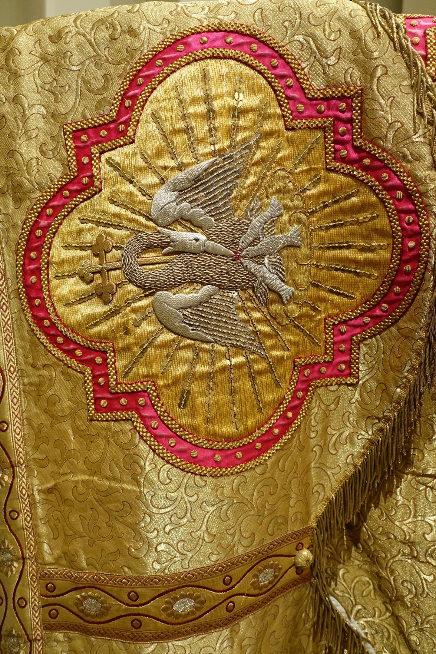 Nahaufnahme eines Priestergewandes mit aufwendiger Gold- und Rotstickerei und einem luxuriösen, königlichen Design vor einer Wand im Hintergrund.