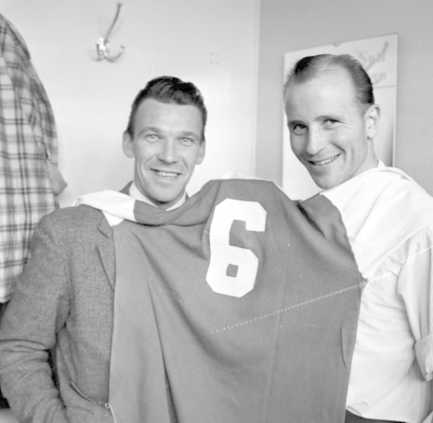 Zwei Männer in Schwarz und Weiß stehen nebeneinander und lächeln, während sie ein Fußballtrikot hochhalten; ein Brett und ein Bügel sind an der Wand hinter ihnen angebracht.