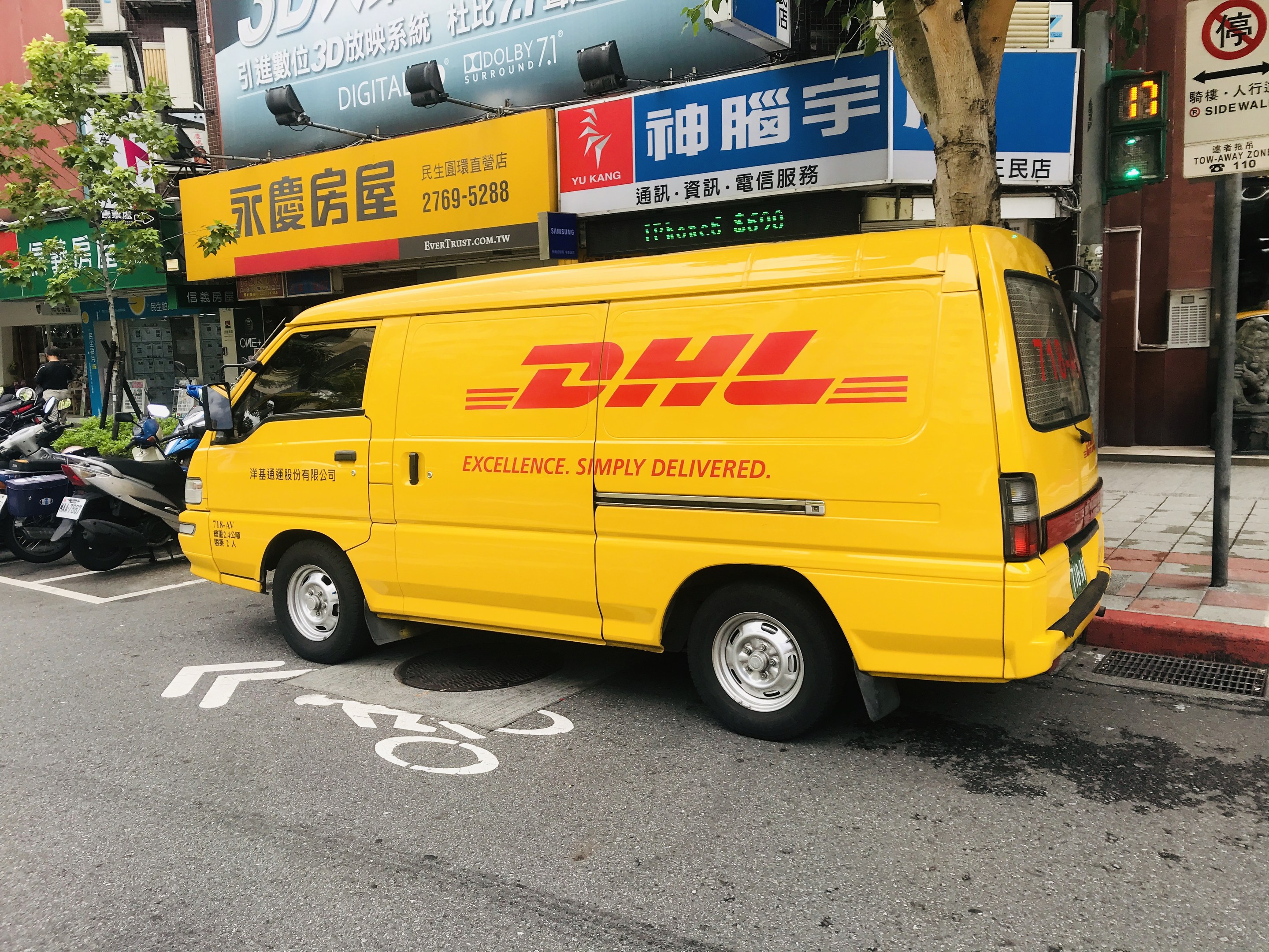 Ein gelber DHL-Lieferwagen parkt auf einer Straße neben Gebäuden, Bäumen, Schildern, Mästängen, Ampeln und einem Fussgänger.