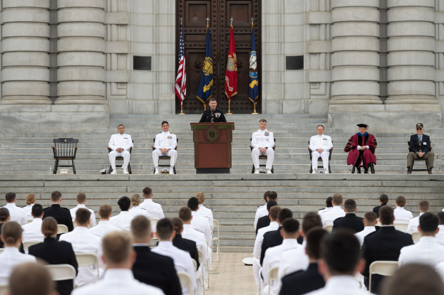 Gruppe von Menschen, die auf Stühlen vor einem Gebäude mit Säulen und einer Tür sitzen, ein Podium mit einem Mikrofon in der Mitte und Fahnen mit Stangen im Hintergrund, was auf eine Abschlussfeier hinweist.