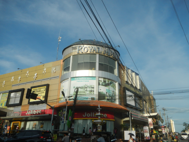 Eine belebte Stadtstraße mit fahrenden Autos, Fußgängern auf dem Gehweg und parkenden Autos vor dem Royal Mall Einkaufszentrum, mit Bäumen, Strommasten und Drähten im Hintergrund unter dem Himmel.