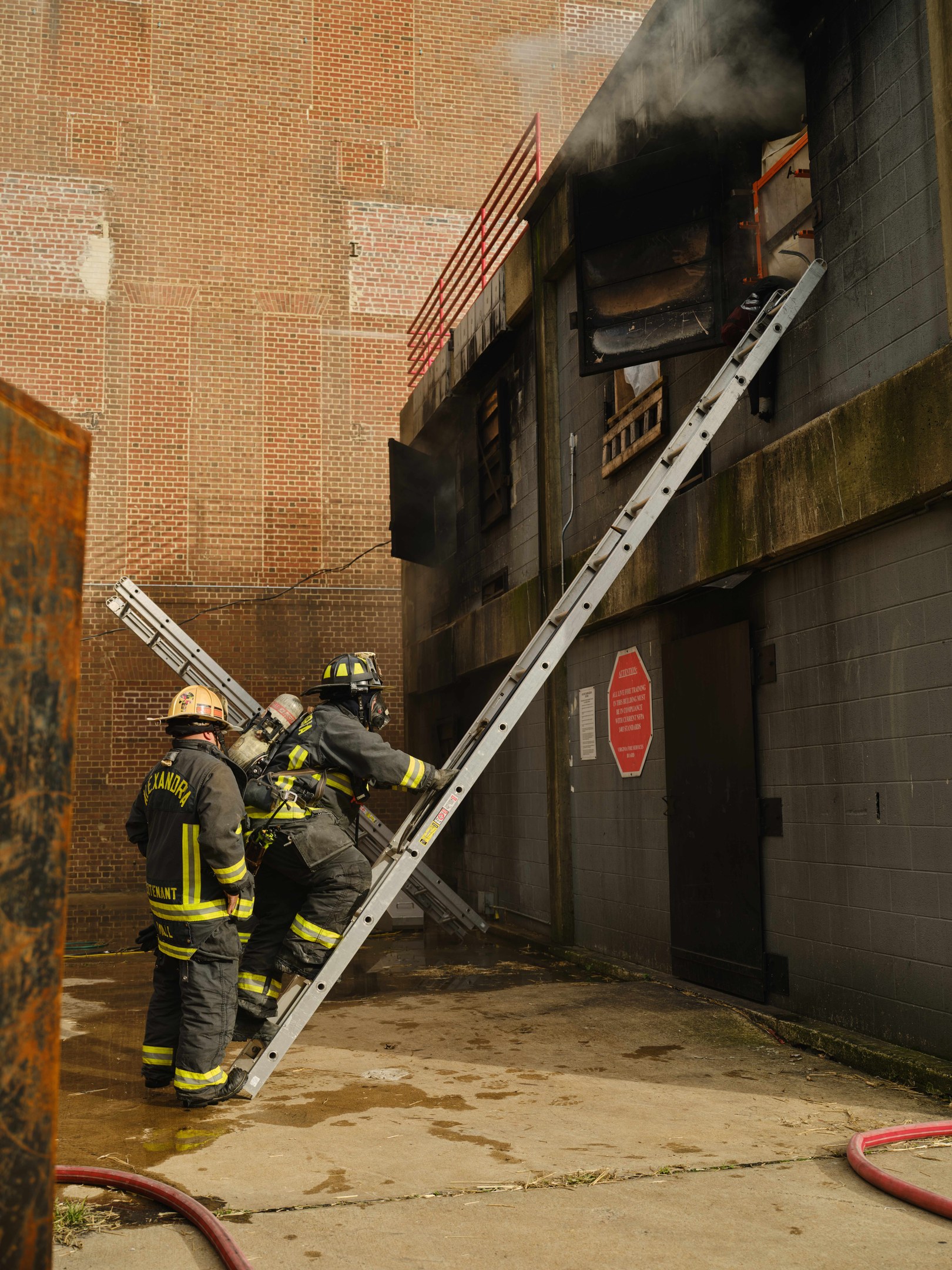 Feuerwehrleute mit Helmen klettern eine Leiter zu einem rauchgefüllten Gebäude hoch, mit einem Metallobjekt links und Rohren auf dem Boden.