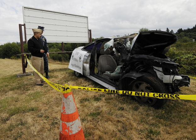 Ein Polizeibeamter in Uniform steht neben einem schwerbeschädigten Auto am Straßenrand, mit eingedrückter Motorhaube und zersplitterten Fenstern, neben einem Verkehrskegel und einem markierten Pfahl, mit Bäumen und einem bewölktem Himmel im Hintergrund.