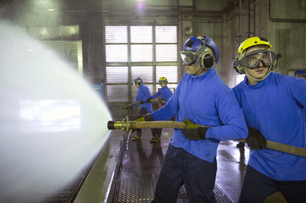 Gruppe von Männern in blauen Hemden und gelben Helmen arbeitet an Maschinen, einer sprüht Wasser auf den Boden in einer Fabrikumgebung mit sichtbaren Rohren, Fenstern und Lichtern.