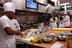 Eine Gruppe von Köchen in einer Küche beim Zubereiten von Speisen auf einem Schneidbrett, wobei ein Koch in Weißem im Vordergrund mit einem Messer in der Hand und umgeben von Utensilien, Schüsseln, Gemüse, einem Mobiltelefon, Flaschen und anderen Gegenständen steht, mit einem Fernseher und Deckenleuchten im Hintergrund.