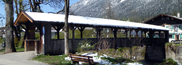 Ein hölzernes Unterschlupf mit schneebedecktem Dach, einer Bank davor, Pflanzen, Gras, einer Gruppe von Bäumen, Häusern mit Dächern und Fenstern, einem Schild, einem Mülleimer, Pfosten, Hügeln und einem bewölkten Himmel.