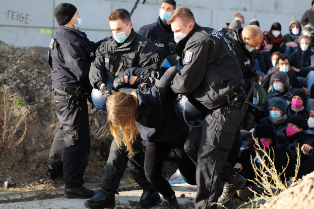 Gruppe maskierter Personen in Mützen, einige bewaffnet, die eine Frau umgeben, die von der Polizei eskortiert wird, mit Pflanzen, Steinen und einer Wand mit Protesttext im Hintergrund.