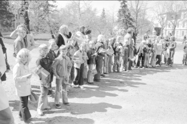 Schwarzes und weißes Bild einer Gruppe von Menschen, die in einer Reihe auf einer Schotterstraße stehen, Fahnen schwingend, mit Bäumen, Gebäuden und einem klaren Himmel im Hintergrund, die an einem Protestmarsch auf dem Schulgelände teilnehmen zu scheinen.