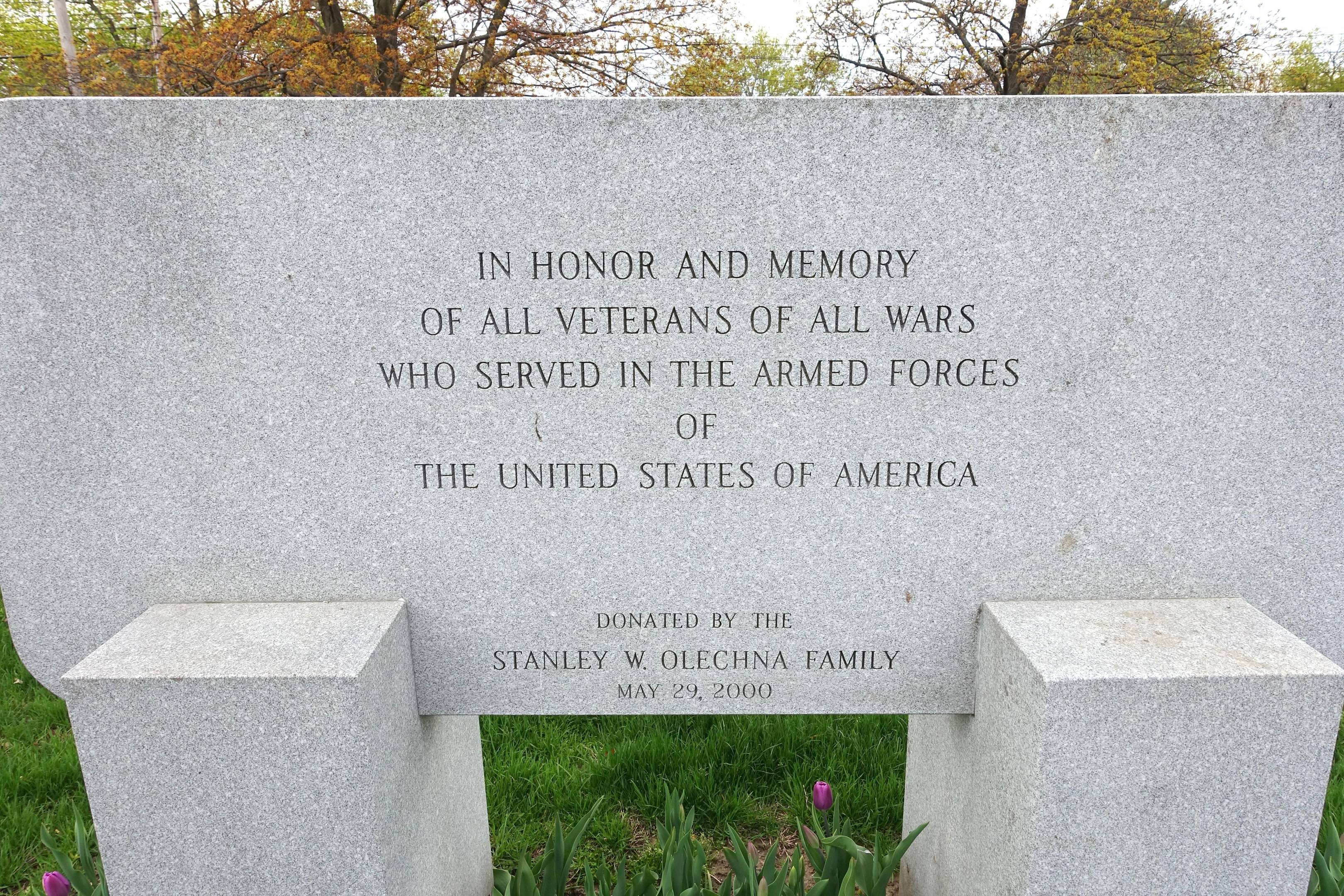 Denkmalstein mit einer Inschrift für US-Armed-Forces-Veteranen, umgeben von Pflanzen, Blumen, Gras, Bäumen und einem bewölkten Himmel.