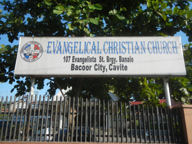 Eine evangelische Kirche in Bacoor City, Cavite, mit einem Schild, einem Metallzaun, einer Wand, Bäumen, Fahrzeugen, Gebäuden, Polen und einem bewölkten Himmel.