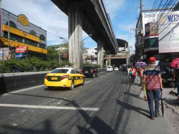 Gelbes Taxi fährt auf einer Straße mit hohen Gebäuden, Menschen mit Regenschirmen auf dem Gehweg, eine Brücke im Hintergrund, bewölkter Himmel und Strommasten mit Drähten entlang der Straße.